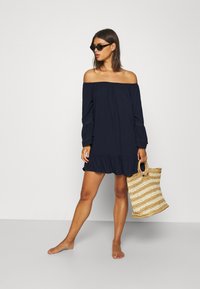 Vestido azul marino con hombros al descubierto, mangas largas y flotantes, y un dobladillo con volantes; acompañado de un bolso de paja trenzada con un patrón a rayas.