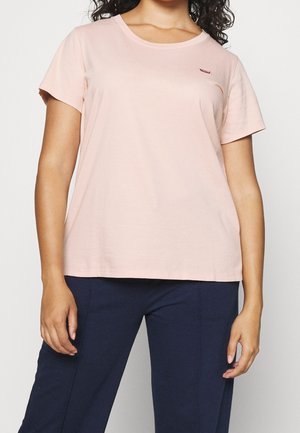 Kvinde iført en lyserød kortærmet t-shirt og mørkeblå bukser, stående mod en ensfarvet hvid baggrund.