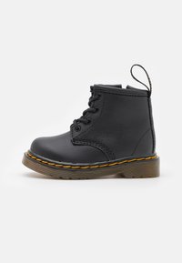 Bottine en cuir noir avec un bout rond, des lacets et une semelle en caoutchouc texturée avec une couture jaune distinctive autour du bord.