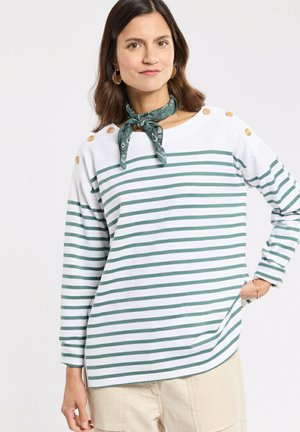 Femme portant un haut à manches longues rayé blanc et vert avec des boutons en bois sur les épaules et un foulard vert à motifs autour du cou, pantalon beige.