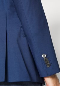 Sacou bleumarin cu o textură netedă, detaliu cu ventilație laterală și patru nasturi negri la manșetă, cu accente de broderie subtile.