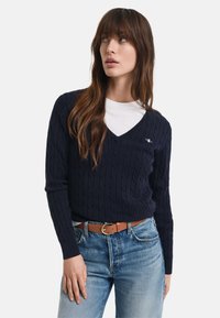 Maritimer Kabelstrickpullover mit V-Ausschnitt und langen Ärmeln, getragen über einem weißen Hemd. Verfügt über ein kleines gesticktes Logo und ein strukturiertes Muster.