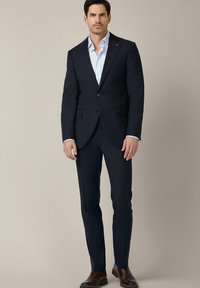 Granatowy garnitur z klapami notch i dwoma guzikami, w połączeniu z jasnoniebieską koszulą. Ciemnobrązowe skórzane buty dopełniają całość.