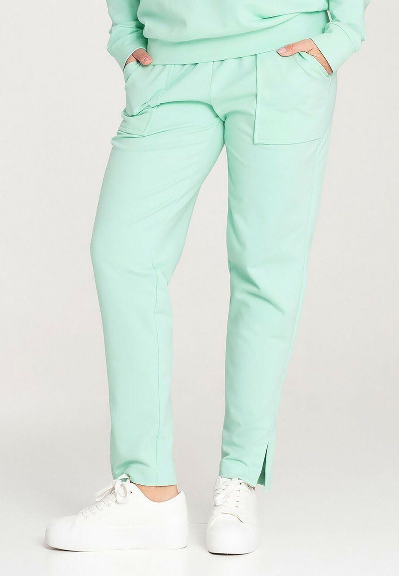Miętowe spodnie o dopasowanym kroju z gładką teksturą, bocznymi kieszeniami i rozcięciami przy kostkach, zestawione z białymi sneakersami.