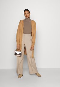 Beige wollen cardigan met capuchon, bruine geribde coltrui, ruime beige broek, gestructureerde instappers en metallic handtas met bovenhandvat.