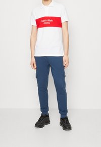 Biała koszulka polo z czerwoną poziomą paską i logo "Calvin Klein Jeans". Granatowe dresowe spodnie cargo z kieszeniami bocznymi i teksturowanym materiałem. Czarne sneakersy.