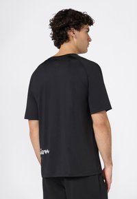 Hombre con cabello oscuro y rizado, vistiendo una camiseta negra de manga corta y pantalones negros, de espaldas contra un fondo blanco.