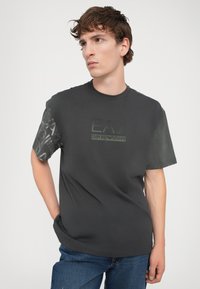 Tamsiai pilka trumpo rankovės marškinėliai. Su tekstūruota raštu ant rankovių ir ryškiu EA7 Emporio Armani logotipu žalios spalvos.