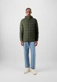 Veste matelassée verte avec capuche, design capitonné et un petit logo jaune sur la poitrine. Portée avec un jean bleu clair et des sneakers beiges.
