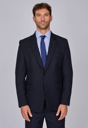 Blazer bleu marine foncé en tissu texturé, dotée de deux poches avant, de revers en pointe et d'une fermeture à un bouton. Porté avec une chemise rayée bleue et une cravate.