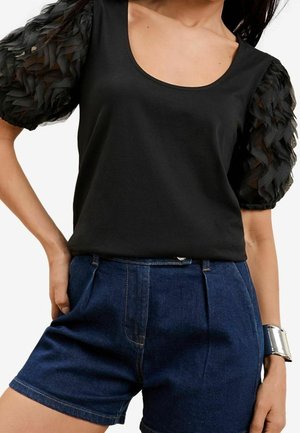 Musta t-paita, jossa on teksturoitu, läpikuultava puffihihna, yhdistettynä korkeavyötäröisiin tummiin farkkushortseihin. Ranteessa hopeinen käsikoru.