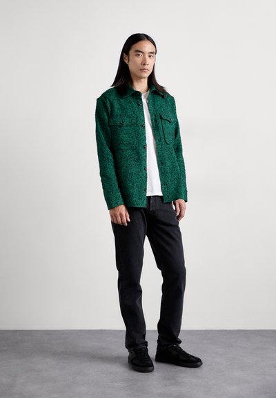 Veste chemise verte texturée avec deux poches sur la poitrine, boutons noirs, associée à un jean noir et des baskets. Design simple et moderne.