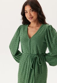 Robe vert en œillets avec un décolleté en V, fermeture à boutons et tissu texturé avec des motifs floraux. Les manches longues et une ceinture à nouer complètent le design.