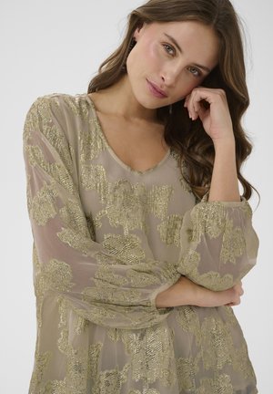 Giovane donna con lunghi capelli castani che indossa una camicetta beige trasparente con motivi floreali dorati, appoggiando la testa sulla mano e guardando verso la fotocamera.