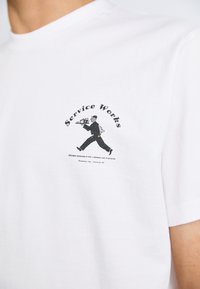 White cotton t-shirt featuring a printed graphic of a figure carrying a tray, with "Service Works" text in a circular layout.