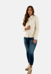Giacca trapuntata bianca con chiusura a bottoni e due tasche, indossata sopra jeans blu e sneakers beige. Presenta un colletto e maniche lunghe.