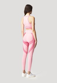 Top sportivo rosa chiaro e leggings con motivi a strisce rosse. I leggings hanno una vita alta. Il modello indossa scarpe da ginnastica bianche. Vista posteriore.