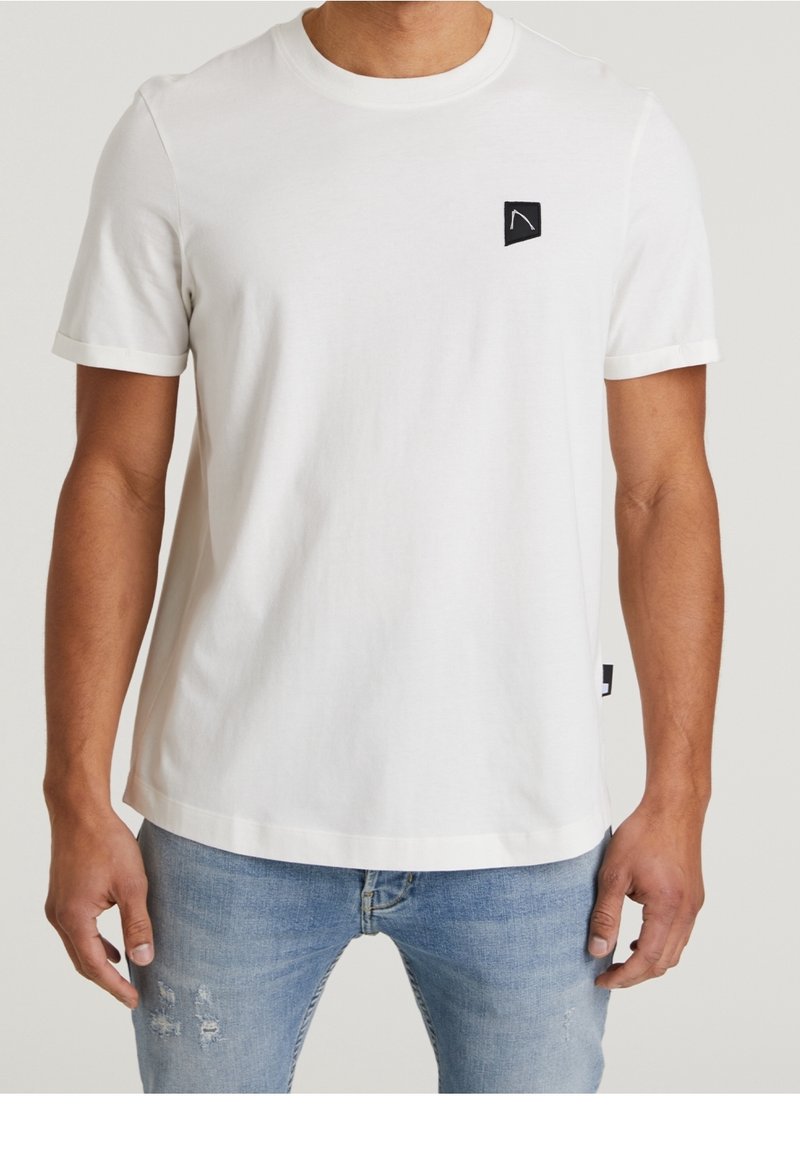 Weißes Baumwoll-T-Shirt mit kurzen Ärmeln, das ein schwarzes Logo-Patch auf der linken Brust und umgeschlagene Ärmel hat. Getragen mit verwaschenen blauen Jeans.