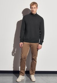 Suéter de cuello alto gris oscuro, de mangas largas, con textura de punto, combinado con chinos beige y zapatillas ligeras, de pie contra un fondo liso.