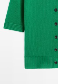 Cardigan en maille verte à manches courtes, bords côtelés et boutons noirs sur fond blanc.