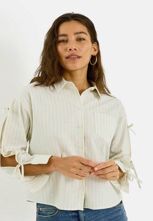 Femme portant une chemise à rayures claires boutonnée avec des manches ouvertes nouées et des boucles d'oreilles anneaux, debout devant un fond uni.