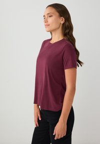 Bordeaux T-shirt met korte mouwen, een ronde hals en een ontspannen pasvorm. Gladde stofstructuur. Gedragen met zwarte broek.