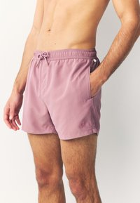 Short de bain pour homme en mauve clair, en tissu lisse. Comprend une ceinture élastique avec cordon de serrage, des poches latérales et un petit logo palmier.
