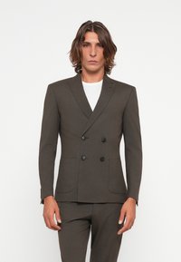 Brown double-breasted suit jacket with lapel, two front pockets, and black buttons. Worn over a white shirt. Textured fabric.