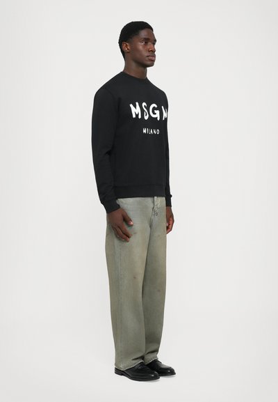 Männlicher Model trägt einen schwarzen MSGM Milano Pullover, lockere hellblaue Jeans mit subtilen Abnutzungsspuren und schwarze Schuhe, steht vor einem schlichten weißen Hintergrund.