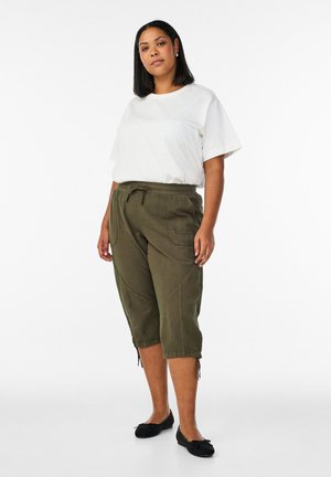 Kvinde står iført en hvid kortærmet skjorte, olivengrønne cropped bukser med snøre og sorte ballet flats, foran en hvid baggrund.