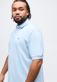 Νεαρός άνδρας με πλεγμένα μαλλιά και γένια, φοράει ανοιχτό μπλε πόλο μπλουζάκι με μικρό λογότυπο σε απλό λευκό φόντο.