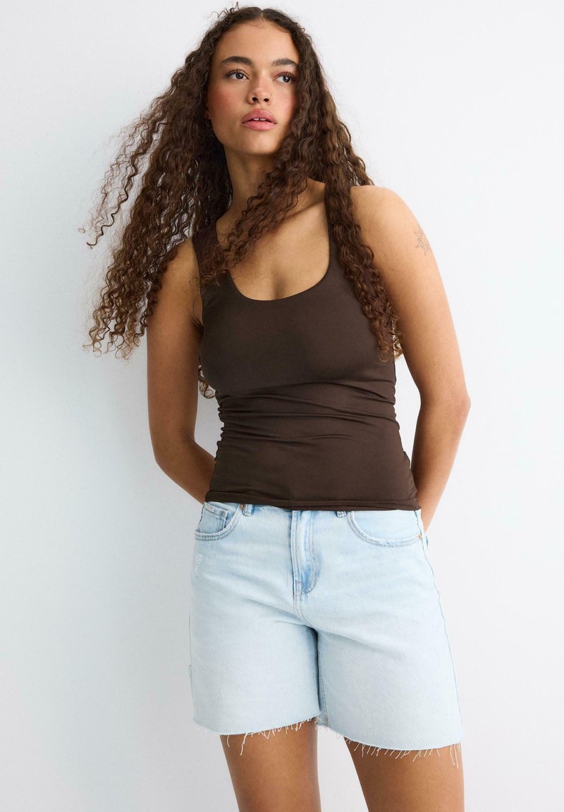 Jeune femme aux cheveux bouclés portant un débardeur marron et un short en jean bleu clair, debout devant un fond blanc.