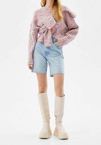 Pull tricot multicolore avec un détail nœud, associé à un short en denim clair et des bottes à plateforme crème montant jusqu'aux genoux. Motif pastel texturé.