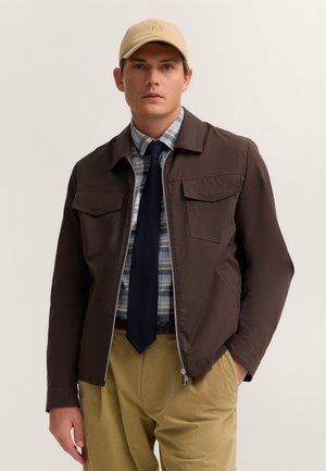 Junger Mann mit beigefarbenem Cap, dunkelbrauner Jacke, blauem Karohemd, schwarzer Krawatte und khakifarbener Hose, stehend vor hellem, einfarbigem Hintergrund.