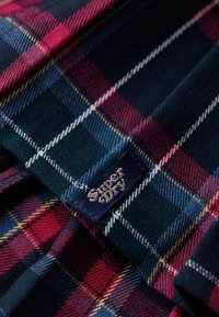 Gros plan sur un tissu à carreaux plié rouge, marine et vert avec une étiquette marine cousue indiquant « Superdry ».