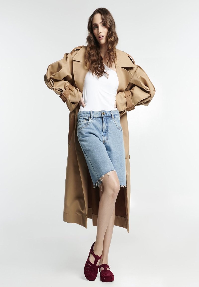 Abrigo trench beige, blusa blanca, pantalones cortos de mezclilla azul claro con dobladillo deshilachado y zapatos burdeos con tiras. Texturas suaves, diseño casual.