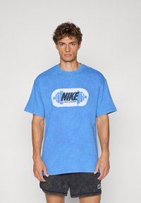 Blå bomulds t-shirt med et fremtrædende sort og hvidt "Nike Athletic Department" logo. Kortærmet og afslappet pasform, båret med grå shorts.