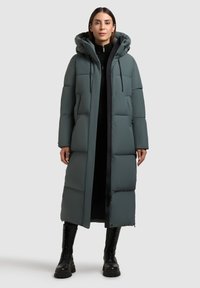 Abrigo largo acolchado en un suave tono teal con capucha, que presenta un patrón acolchado, detalles en negro y cierre de cremallera. Modelo combinado con botas negras.