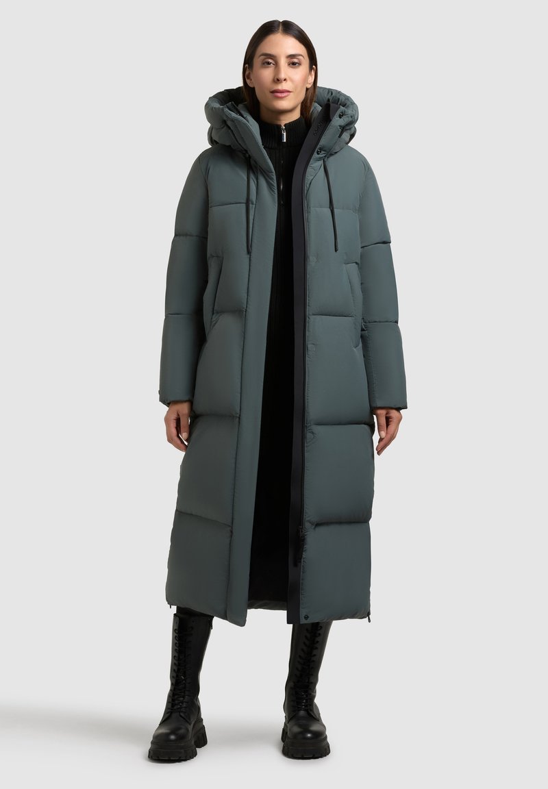 Abrigo largo acolchado en un suave tono teal con capucha, que presenta un patrón acolchado, detalles en negro y cierre de cremallera. Modelo combinado con botas negras.