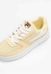 Baskets FILA beiges en cuir perforé, lacets blancs, semelle blanche et logo sur la languette et le côté, inclinées pour montrer le dessus et le côté extérieur.