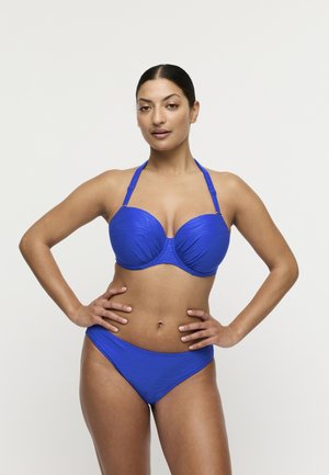 Vrouw poseert met handen in haar zij, draagt een gestructureerde blauwe bikini met halternekbanden tegen een effen witte achtergrond.