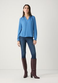 Blausa mit langen Ärmeln in Blau und V-Ausschnitt, kombiniert mit schmalen Jeans in dunklem Denim. Das Model trägt hohe bordeauxfarbene Lederstiefel mit Blockabsätzen.