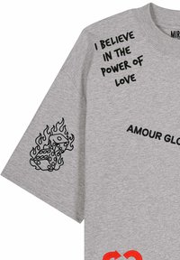 T-shirt gris avec texte noir "I BELIEVE IN THE POWER OF LOVE," texte partiellement visible "AMOUR GLO" et graphique noir de dés enflammés sur la manche.