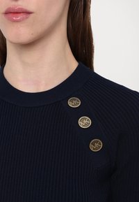 Mörkblå rätstickad tröja med rund halsringning och tre logoknappar i guldton på axeln. Har en mjuk textur och en figurnära design.