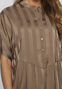 Abito in raso marrone con strisce verticali, colletto alla mandarino e chiusura con bottoni decorativi e pieghe morbide.
