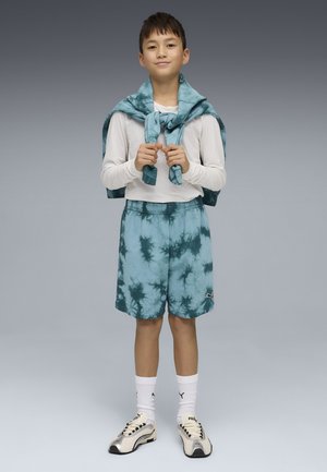 Niño de pie, vestido con camisa blanca de manga larga, pantalones cortos azul teñidos, sudadera a juego atada alrededor de los hombros, calcetines blancos y zapatillas beige.