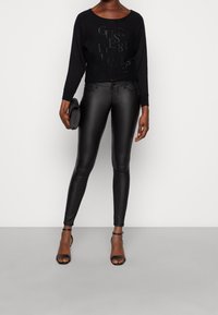 Suéter negro corto con logotipo en relieve, combinado con pantalones ajustados de imitación cuero negros y tacones negros con tiras. Sosteniendo un bolso de mano negro.