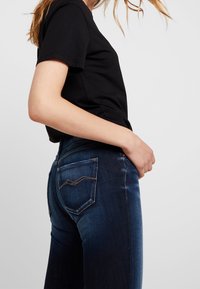 Vaqueros de mezclilla azul oscuro con un ligero desvanecimiento, con bolsillos traseros cosidos y un diseño clásico de cinco bolsillos. Combinados con una camiseta negra.