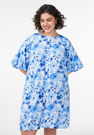 Kvinde med kort krøllet hår iført en løs, knælængde hvid kjole med blå blomsterprint, smilende og kigger ned.