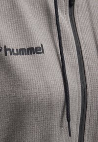 Graue, gerippte Zip-Jacke mit einem schwarzen Reißverschluss und Kordelzug, versehen mit einem mattschwarzen Logo im Brustbereich.